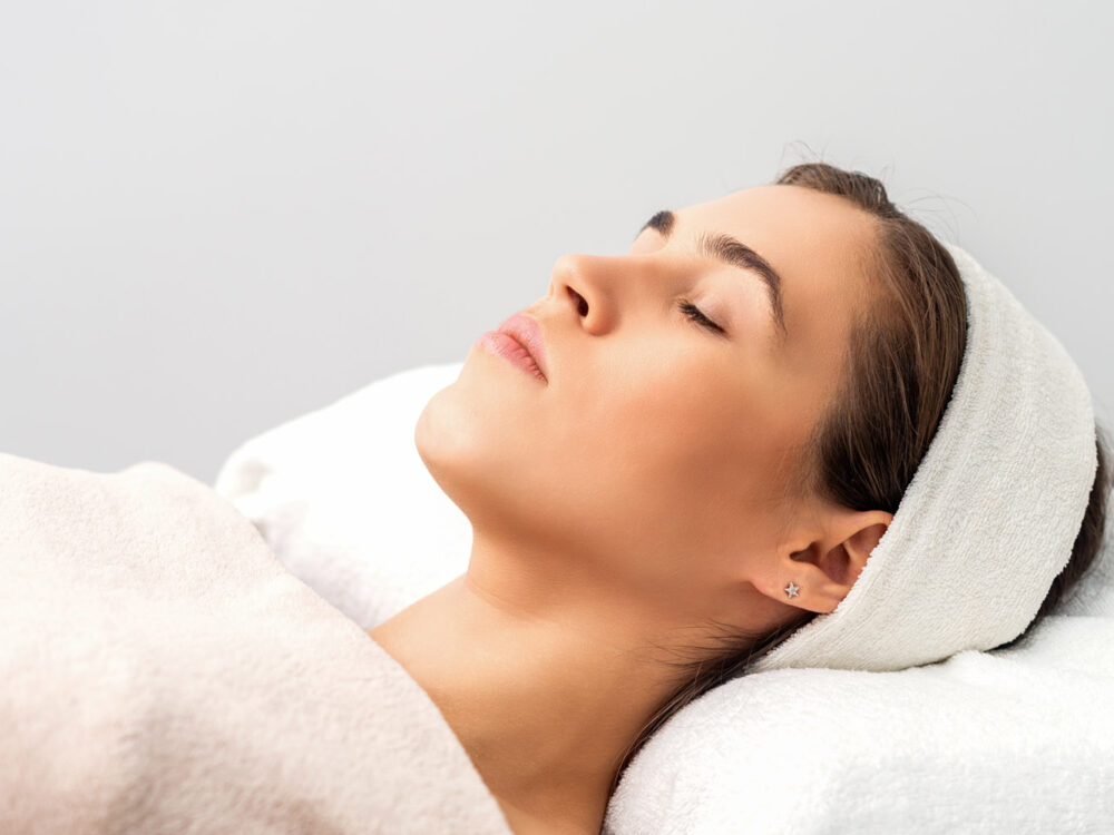 Side view of young woman lying on beautician table with closed eyes while waiting for cosmetic procedure in beauty salon